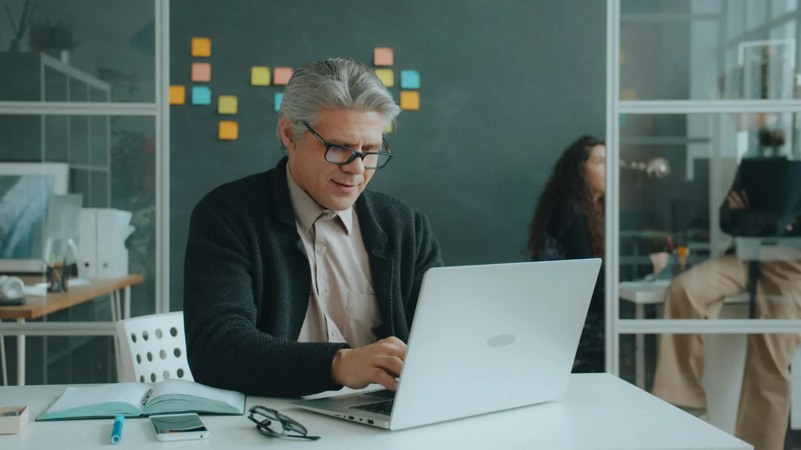 A grey-haired man smiles at his laptop in a modern office while a colleague works behind him