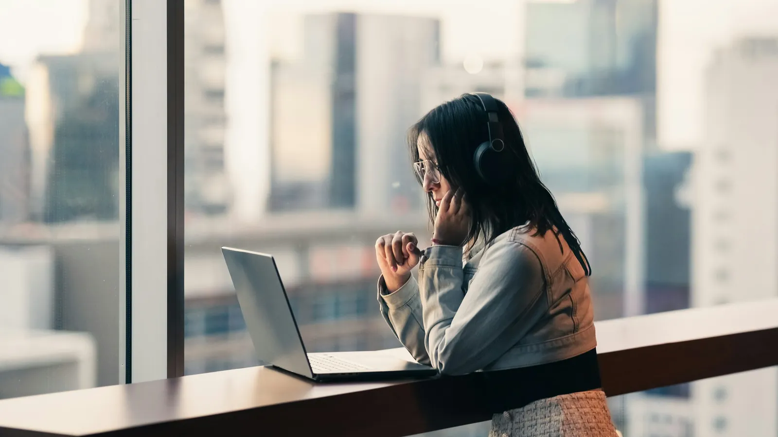 A woman in headphones works alone at a laptop beside a tall window overlooking the city