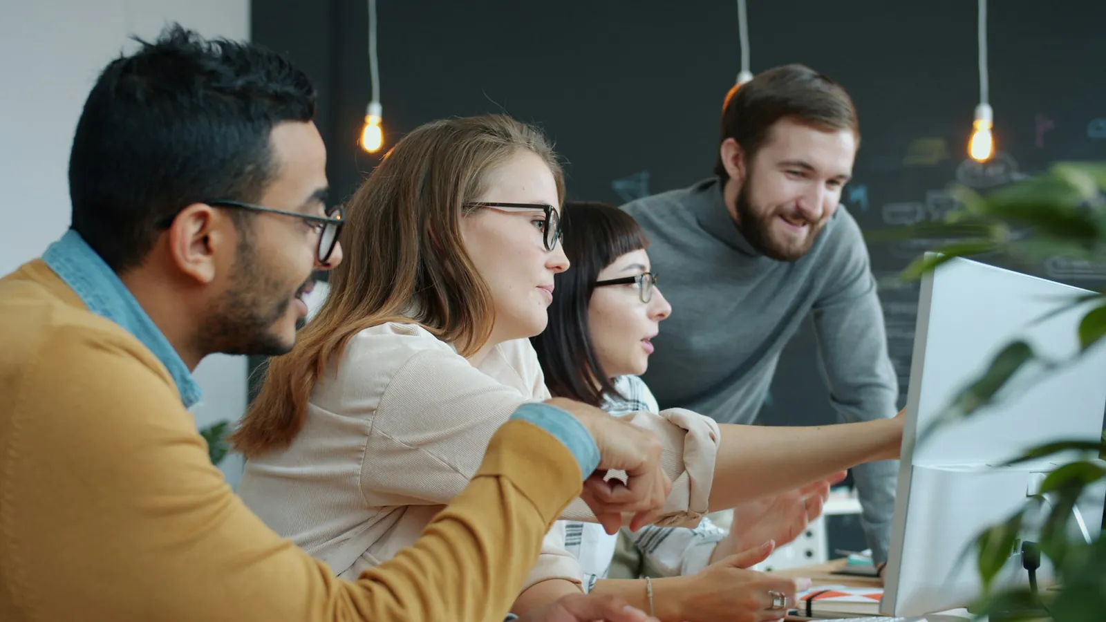 Four colleagues gather around a monitor in a startup office, smiling and pointing at the screen