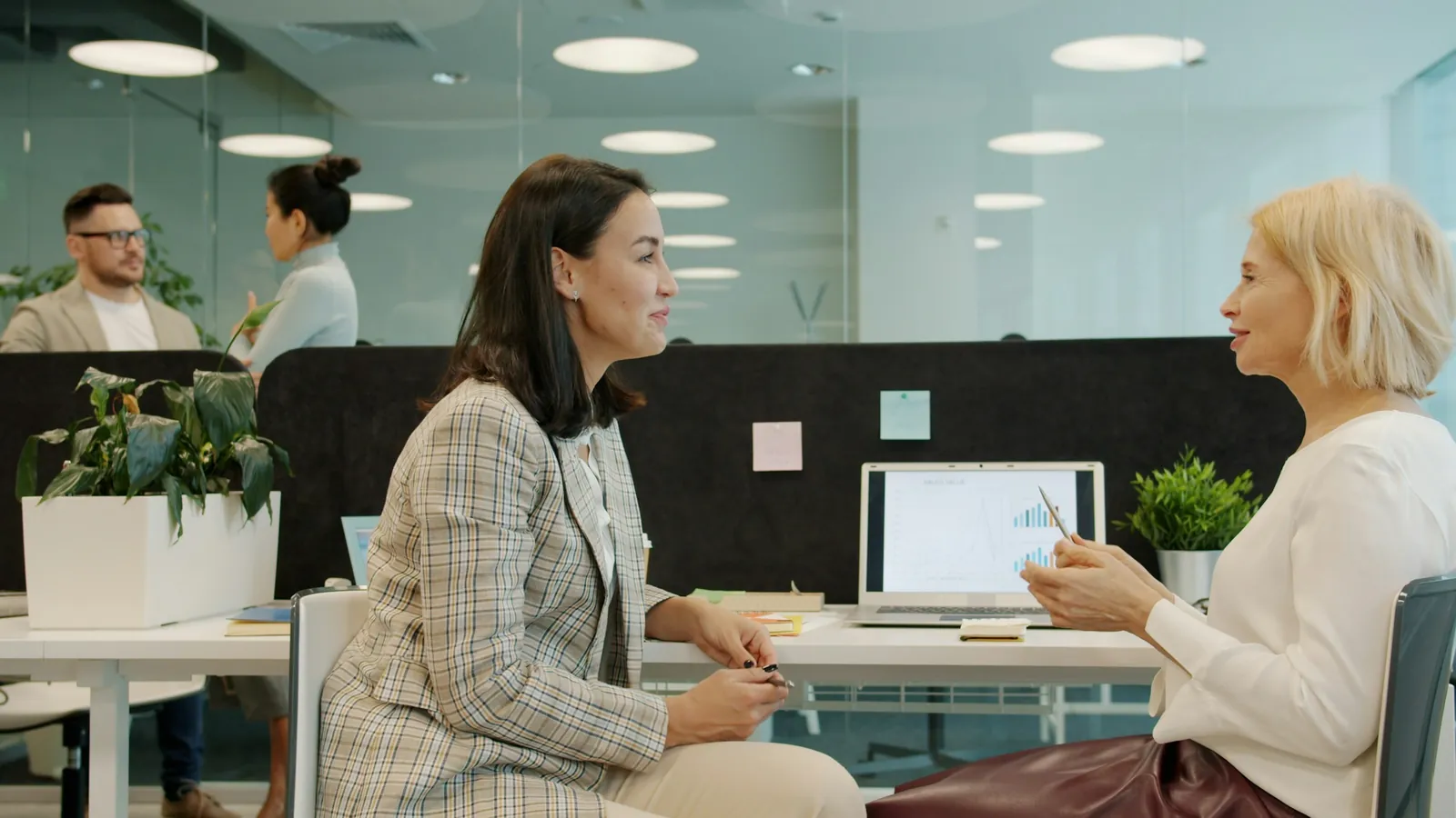 Two women in conversation across a desk in a bright open-plan office, colleagues blurred behind them