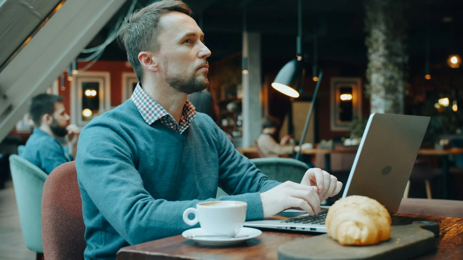 A man in a blue sweater works on a laptop at a cafe table, looking up in thought