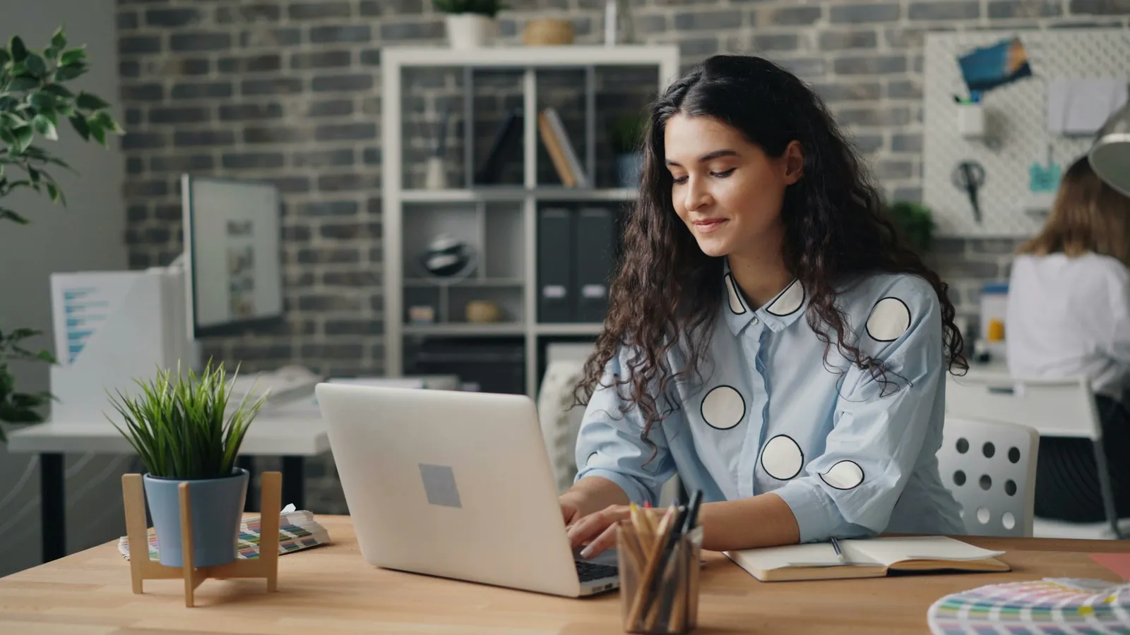 A young woman with curly hair smiles while typing on a laptop at her office desk
